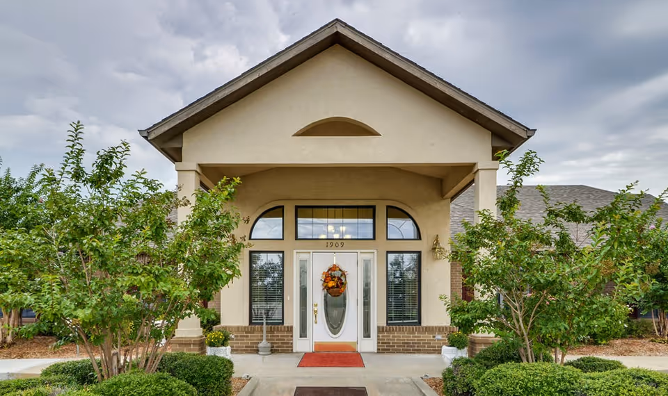 Front exterior view of a senior living facility entrance with a peaked roof, large windows, a white door decorated with a fall-themed wreath, and surrounded by green shrubs and trees under a cloudy sky.