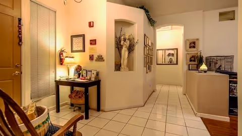Interior view of a warmly lit hallway in a senior living facility with tiled floors, a small table with a lamp and decorative items, framed pictures on the walls, and a niche with a floral arrangement. There is a rocking chair and a basket near the entrance door, and a partial view of a living area with a couch and pet cage.