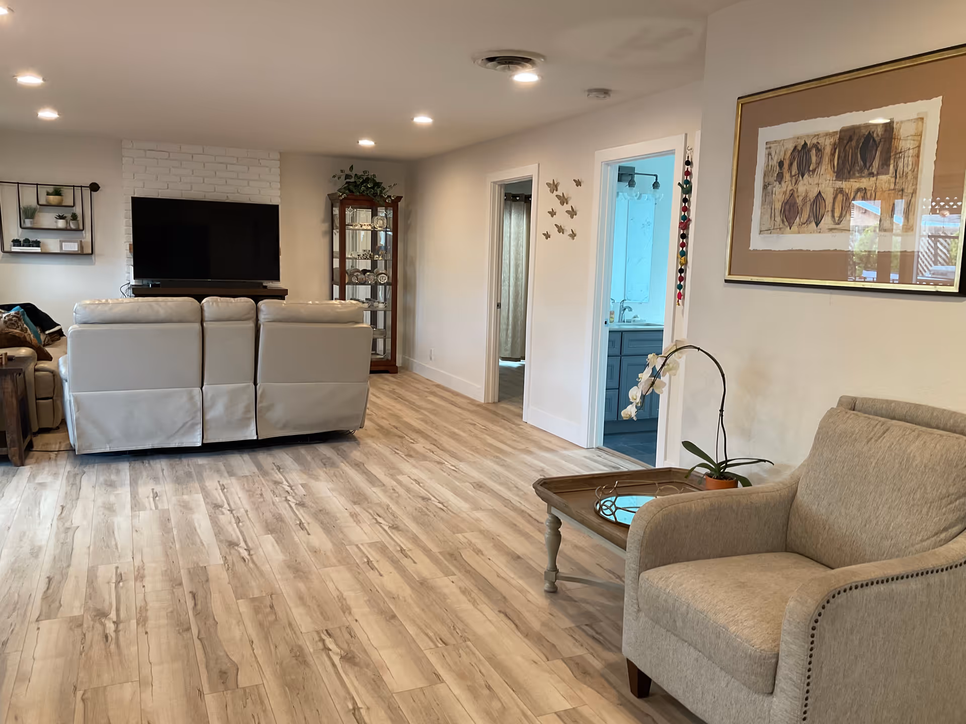 A spacious living room with light wood flooring, a beige armchair with a small wooden coffee table holding a potted orchid, a large flat-screen TV mounted on a white brick wall, a beige leather sofa facing the TV, a glass display cabinet with decorative items, and two doorways leading to other rooms. The walls are decorated with framed artwork and butterfly wall decals.