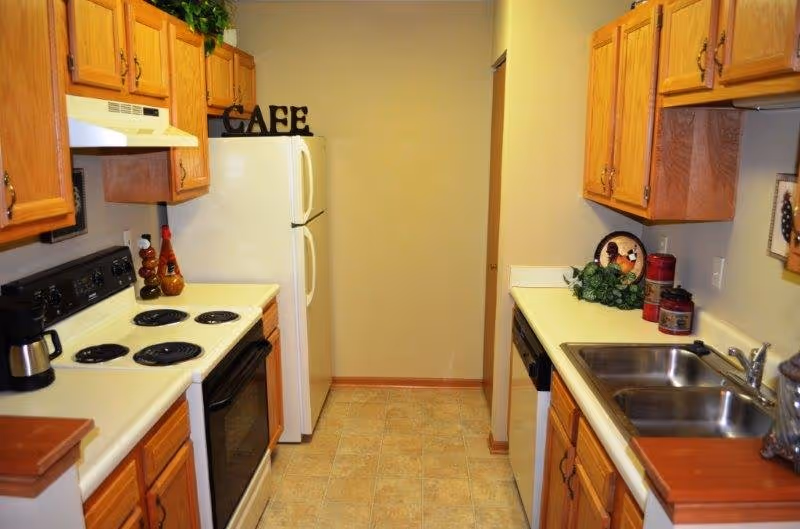 A narrow kitchen with wooden cabinets on both sides, a white refrigerator at the end, a white electric stove with four burners and an oven, a coffee maker on the counter, a double stainless steel sink, and decorative items including a plant and jars. The word 'CAFE' is displayed on top of the refrigerator.