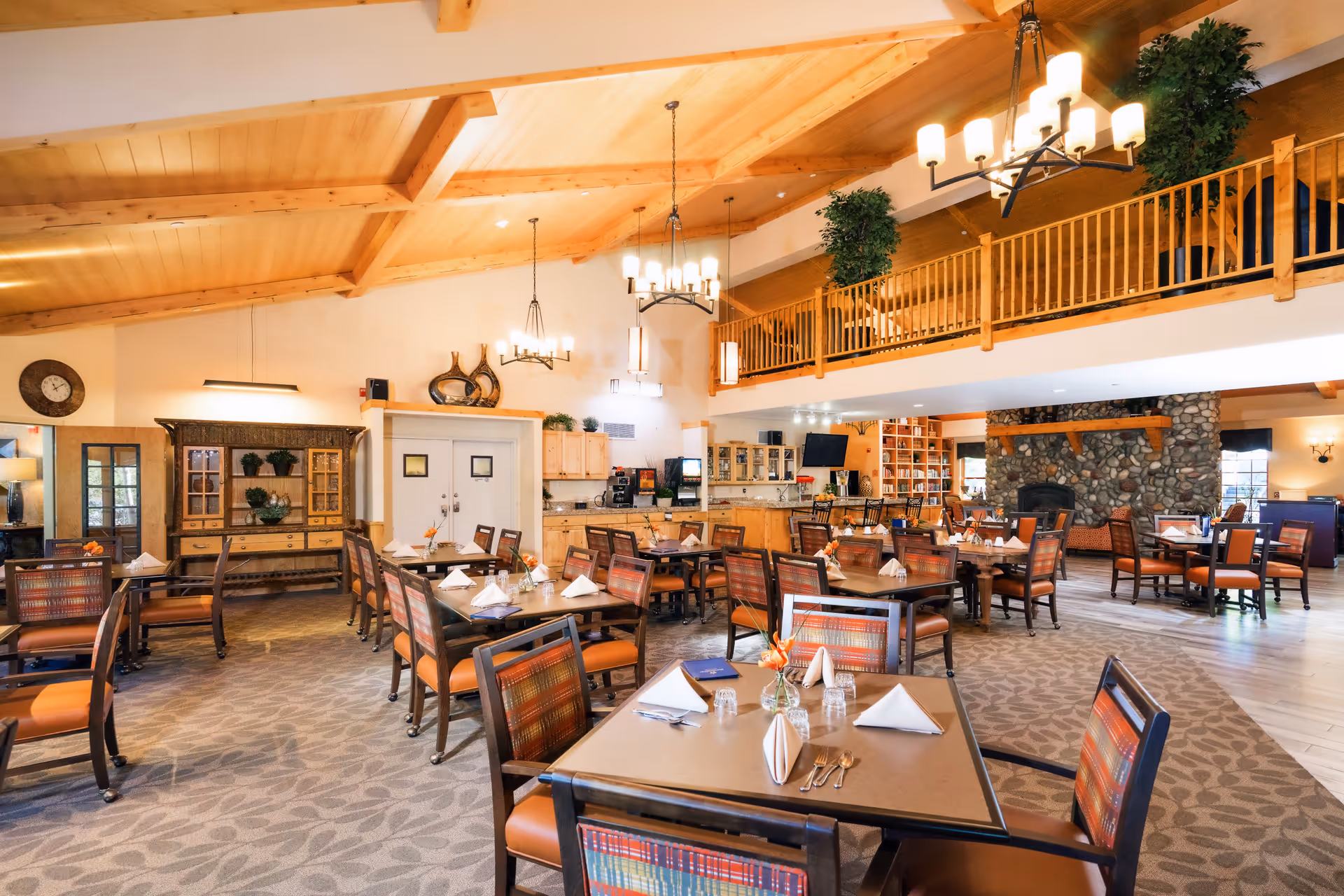 A spacious dining room with multiple tables and chairs arranged neatly. Each table is set with folded napkins, glasses, and silverware. The room features a high wooden ceiling with exposed beams, several hanging chandeliers, and large windows allowing natural light. There is a stone fireplace in the background and a balcony with plants above. The decor includes wooden furniture and warm tones, creating a cozy and inviting atmosphere.