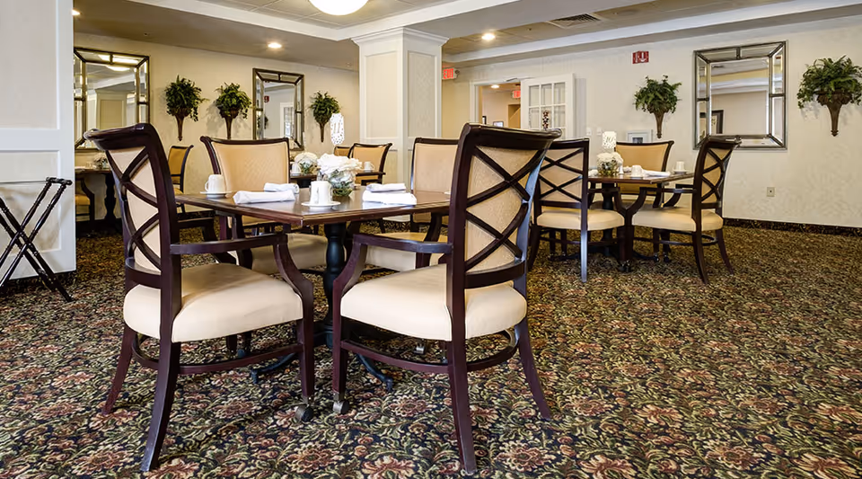 A dining room with several wooden tables and cushioned chairs arranged neatly. Each table is set with white napkins, cups, and small flower arrangements. The room has patterned carpet flooring, light-colored walls, mirrors, and decorative plants mounted on the walls.