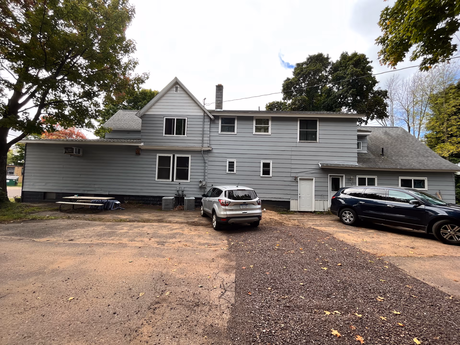 Gray two-story residential building with a small parking area, two parked cars, and surrounding trees.