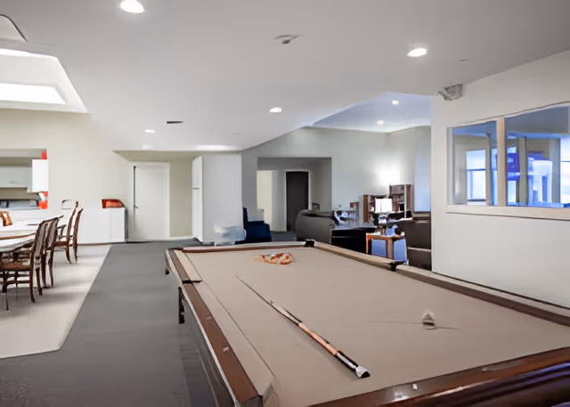 Interior view of a senior living facility common area featuring a pool table in the foreground with pool cues and balls arranged on it. To the left, there is a dining area with a table and chairs. In the background, there is a lounge area with sofas and bookshelves. The room is well-lit with recessed ceiling lights and has a neutral color scheme.