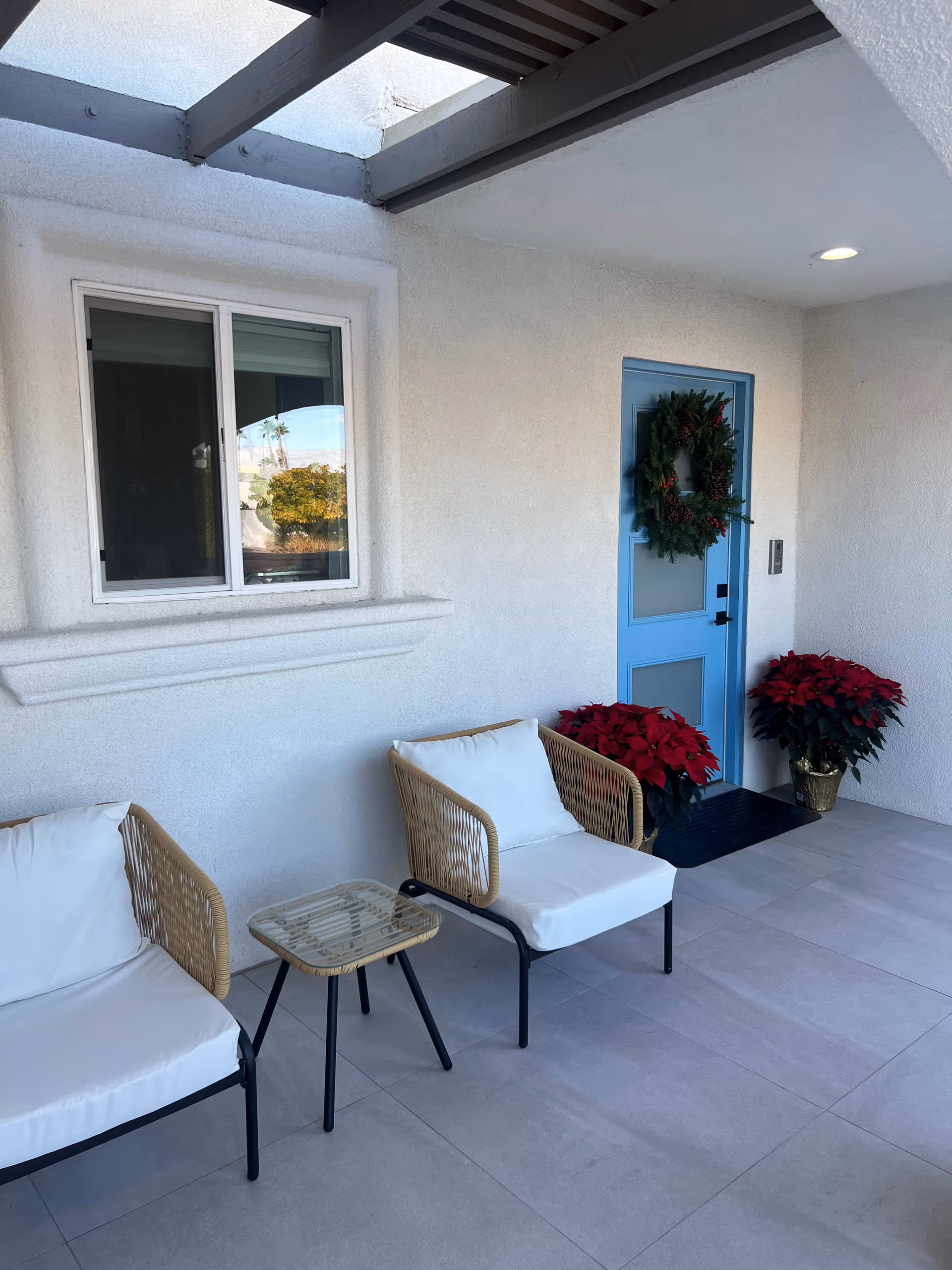 Covered entrance patio with two cushioned chairs and small table beside a blue door decorated with a wreath and poinsettias.