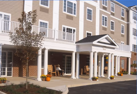 Exterior view of a senior living facility named Parkview at South Pantops showing a multi-story building with beige siding and white trim. The entrance features white columns supporting a small portico, with rocking chairs on the porch and potted plants including pumpkins near the entrance. A tree and landscaped area are visible in front of the building.