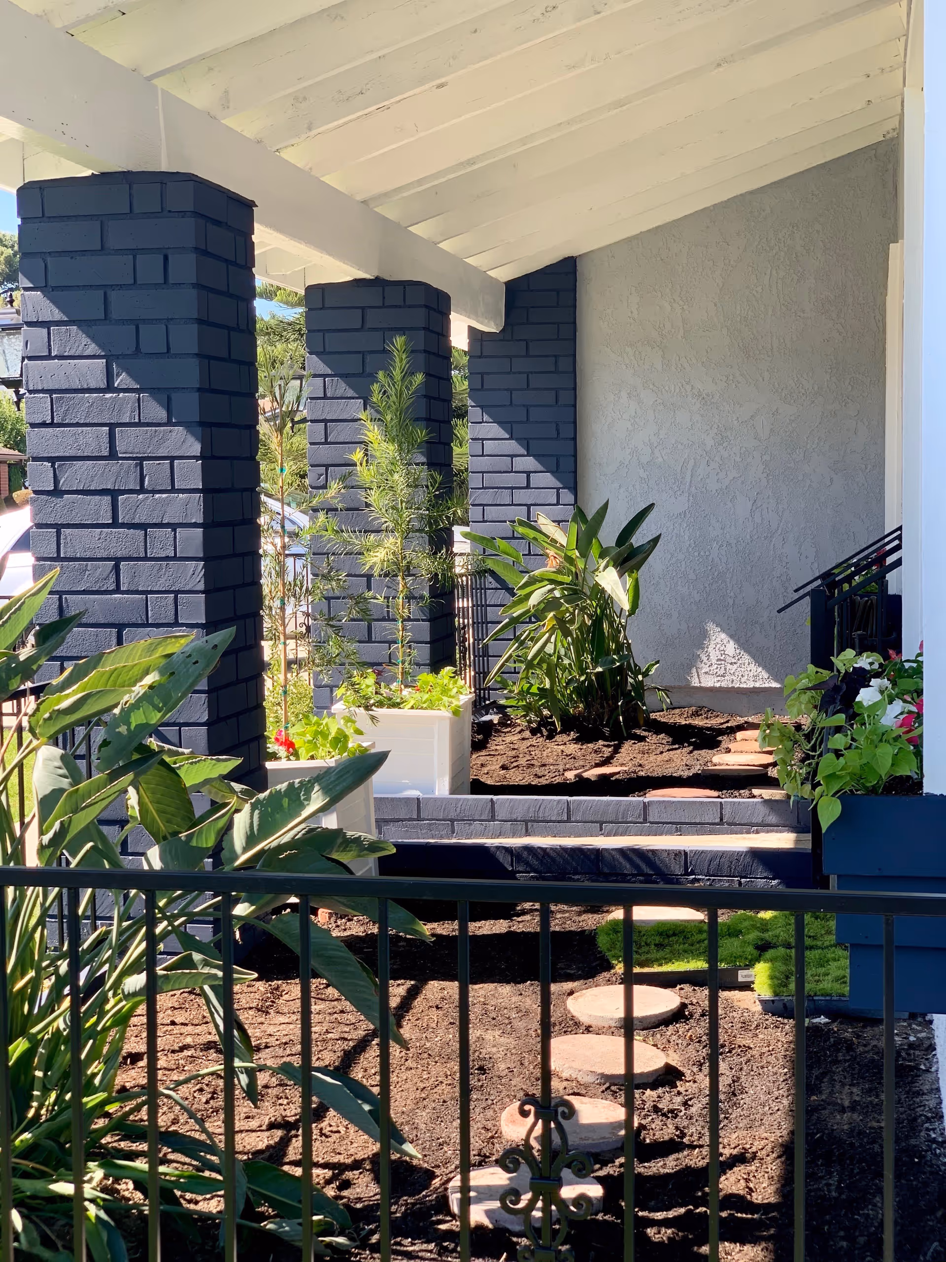 A covered porch area with dark blue brick pillars and a white ceiling. There are various green plants and flowers in planters and garden beds, with a stone stepping path leading through the soil. A black metal fence is visible in the foreground.