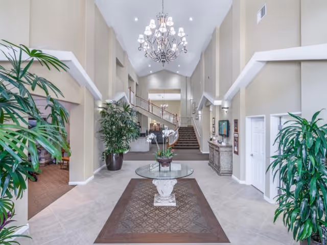 Spacious and well-lit interior lobby area with high vaulted ceilings, a large chandelier, a round glass table with a floral arrangement in the center, potted plants on either side, and a staircase leading to an upper level in the background.