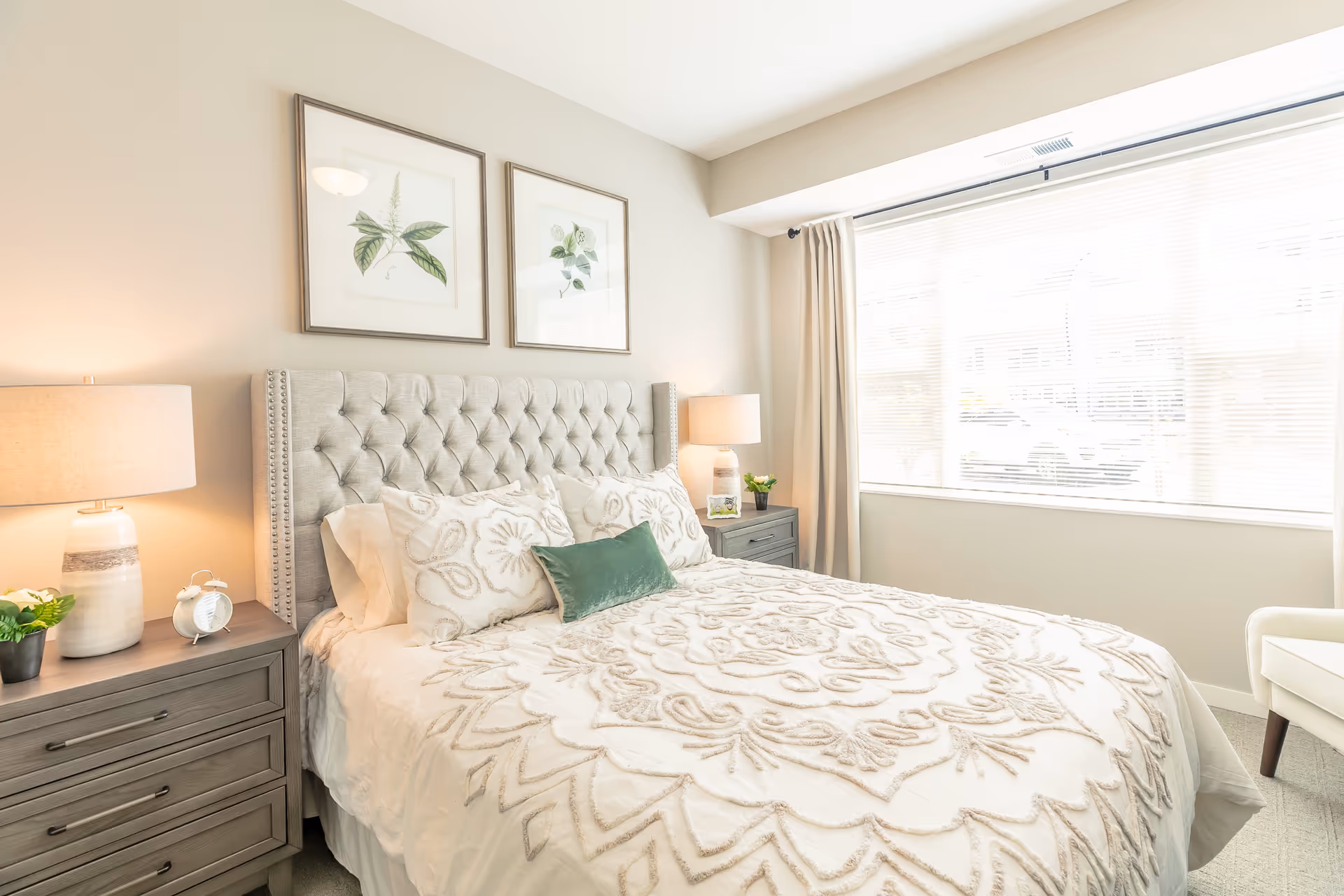A bright and cozy bedroom with a large bed featuring a tufted headboard and decorative white bedding with a green accent pillow. There are two matching nightstands on either side of the bed, each with a lamp and small decorative items. Two framed botanical prints hang on the wall above the headboard. A large window with blinds and curtains allows natural light to fill the room.
