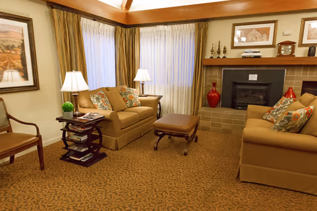 Living room with two beige sofas, an ottoman, side tables with lamps, patterned carpet, and a fireplace.