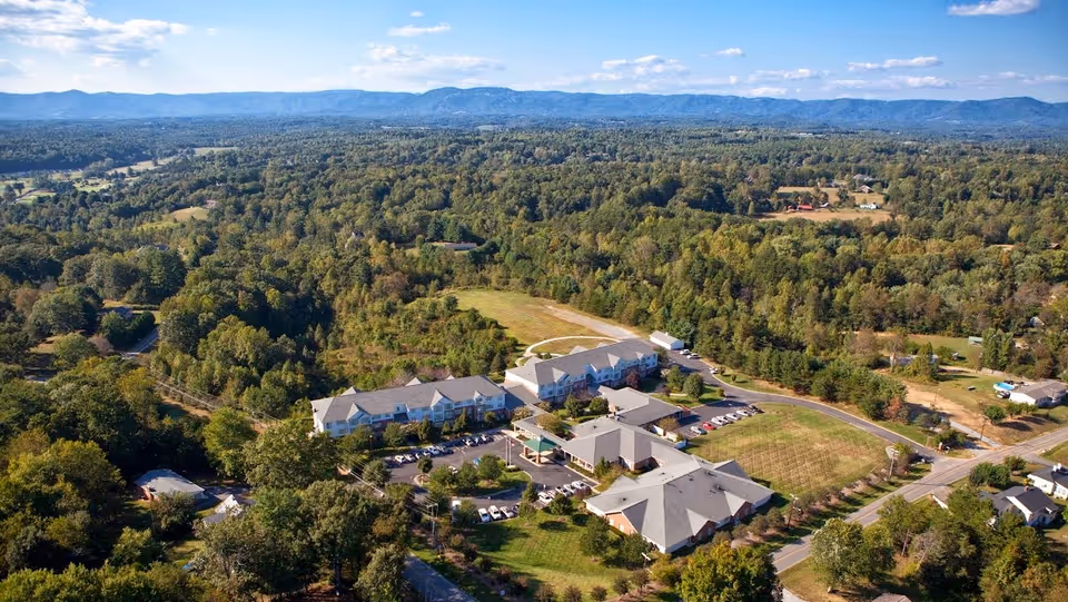 Aerial view of RidgeCrest senior living facility surrounded by dense green trees and open fields, with a clear blue sky and distant mountains in the background.