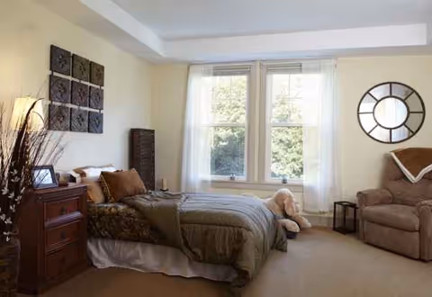 A cozy bedroom in Sunrise of Weston with a bed, window, seating area, and decor.