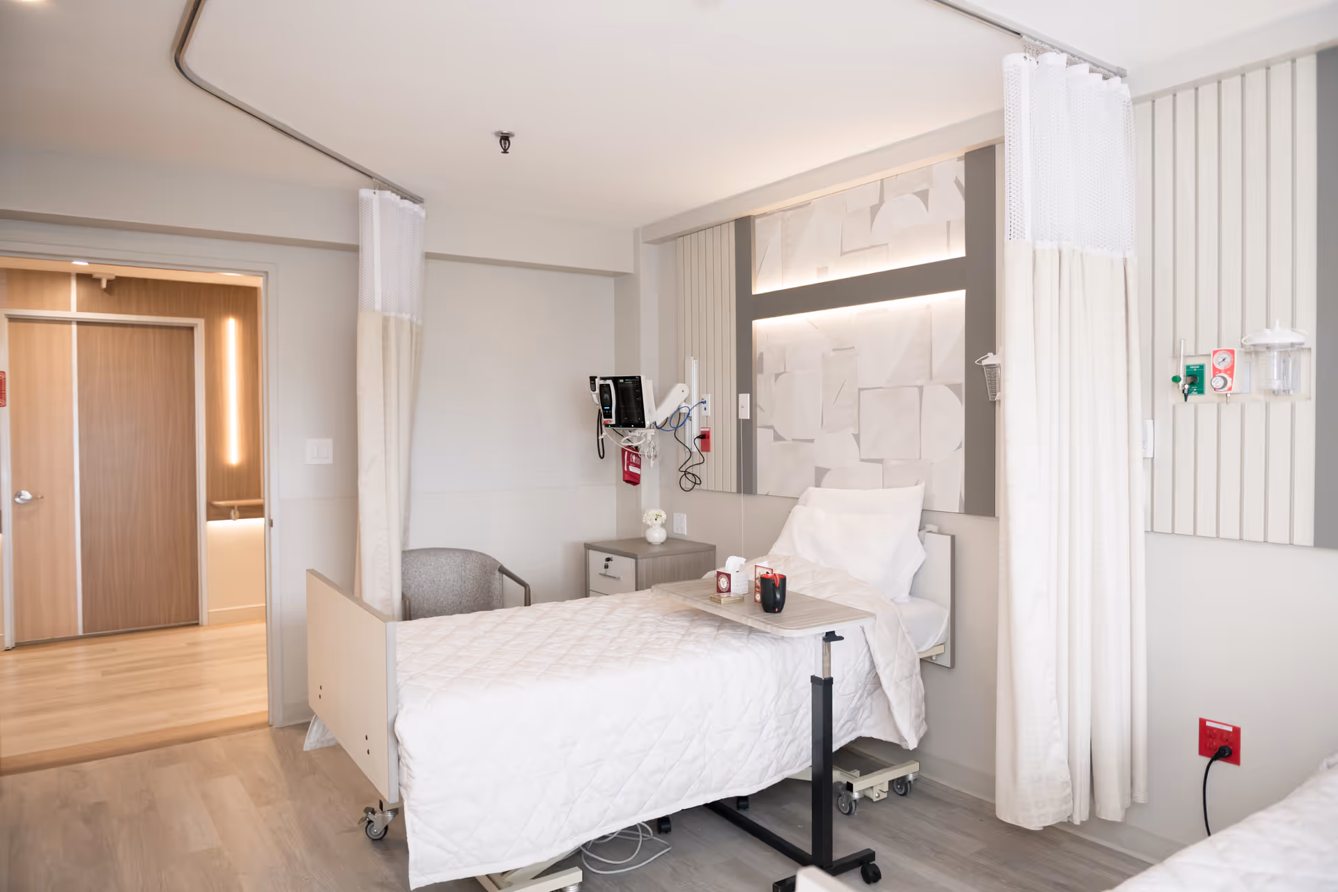 A bright private skilled nursing room with a hospital bed, over-bed table, medical monitors, privacy curtains, and a chair.