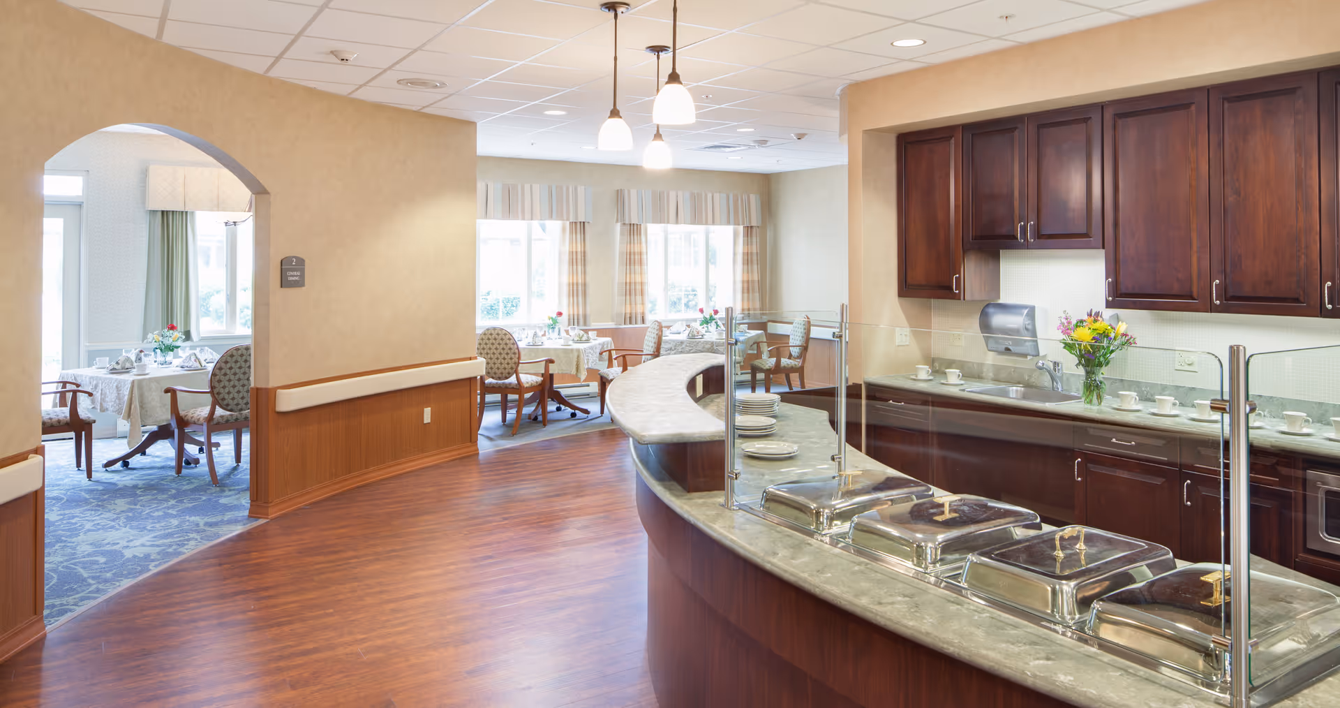 Interior view of a senior living facility dining area with a curved serving counter featuring covered food trays and a vase of flowers. In the background, there are tables set with tablecloths, chairs, and place settings near large windows with curtains, allowing natural light to fill the room.