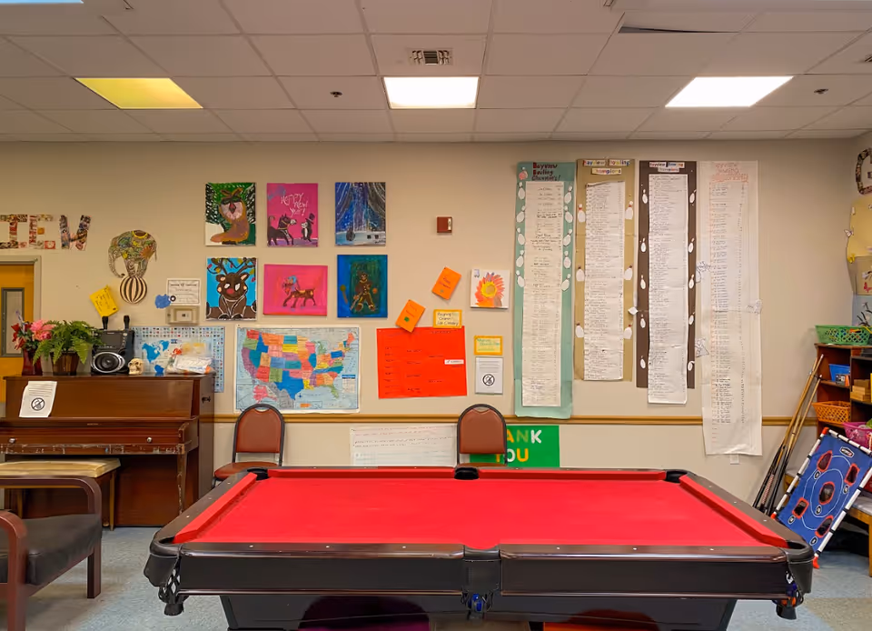 A recreational room with a red felt pool table in the center. Behind the pool table, there are two chairs against a wall decorated with colorful paintings, a map of the United States, various charts, and notes. To the left, there is a piano with a plant and a radio on top. The ceiling has fluorescent lights.