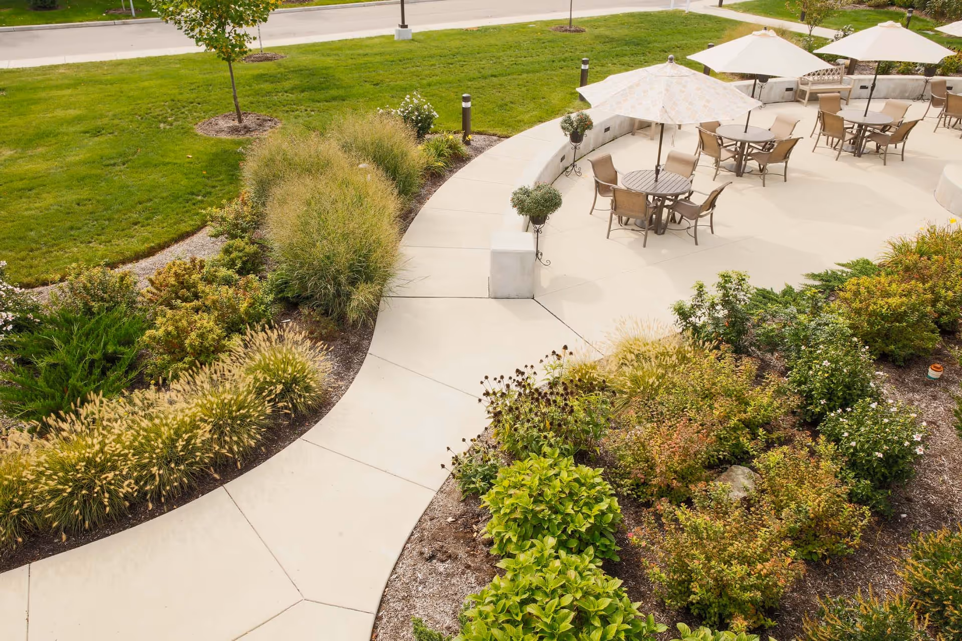 Curved paved walkway leading to an outdoor patio with tables, chairs, umbrellas, and surrounding landscaped gardens.