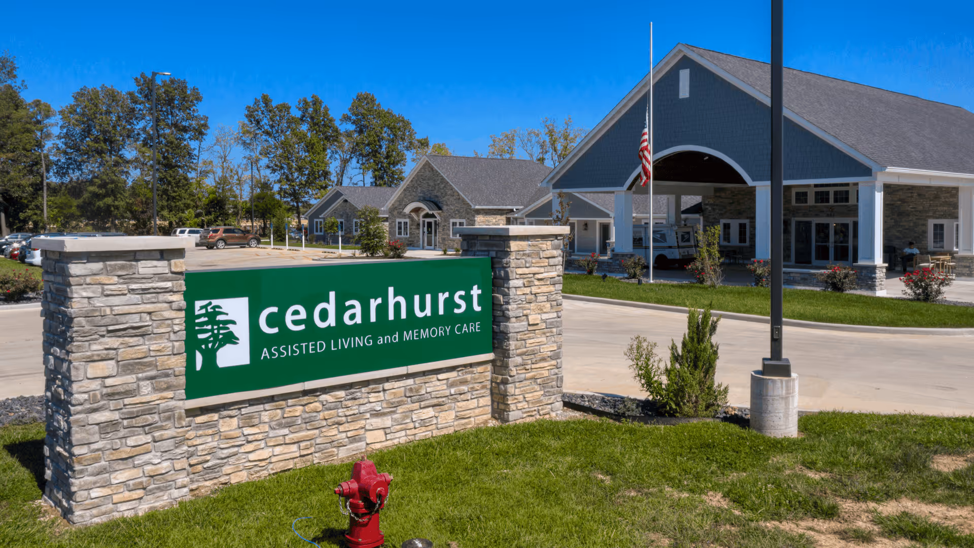 Exterior view of Cedarhurst assisted living and memory care facility with a stone sign in the foreground displaying the facility name and logo. The building has a covered entrance, stone and siding exterior, and an American flag at half-mast. There are trees and parked cars in the background under a clear blue sky.