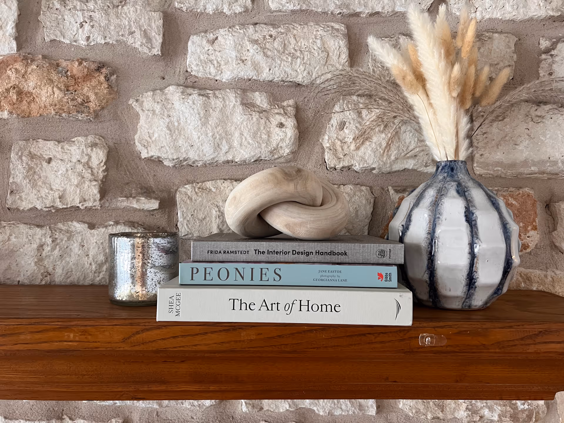 A wooden shelf mounted on a stone wall holding three stacked books titled 'The Interior Design Handbook', 'Peonies', and 'The Art of Home'. On top of the books is a decorative wooden knot sculpture. To the right of the books is a white and blue ceramic vase with dried pampas grass, and to the left is a small metallic candle holder.