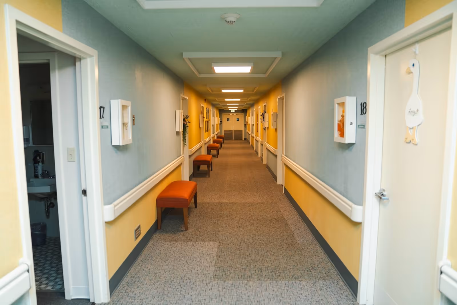 A long, well-lit hallway in a senior living facility with yellow and light blue walls, carpeted floor, handrails on both sides, and several orange cushioned benches along the walls. Doors to rooms line both sides of the hallway, with one door open showing a bathroom inside.