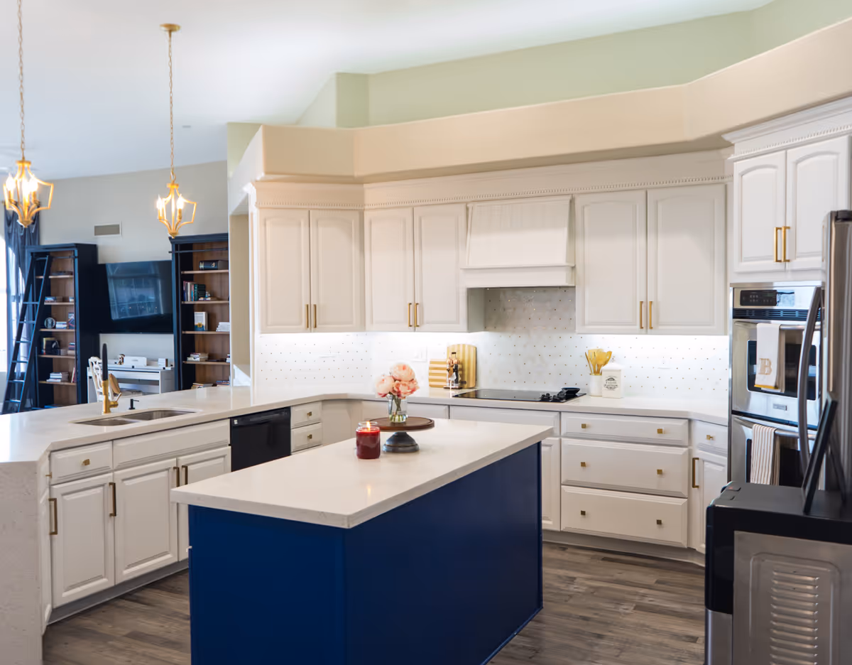Bright and modern kitchen with white cabinetry, a blue island with a white countertop, two pendant lights hanging above the island, stainless steel appliances including a refrigerator and oven, and a living room area visible in the background with bookshelves and a wall-mounted TV.