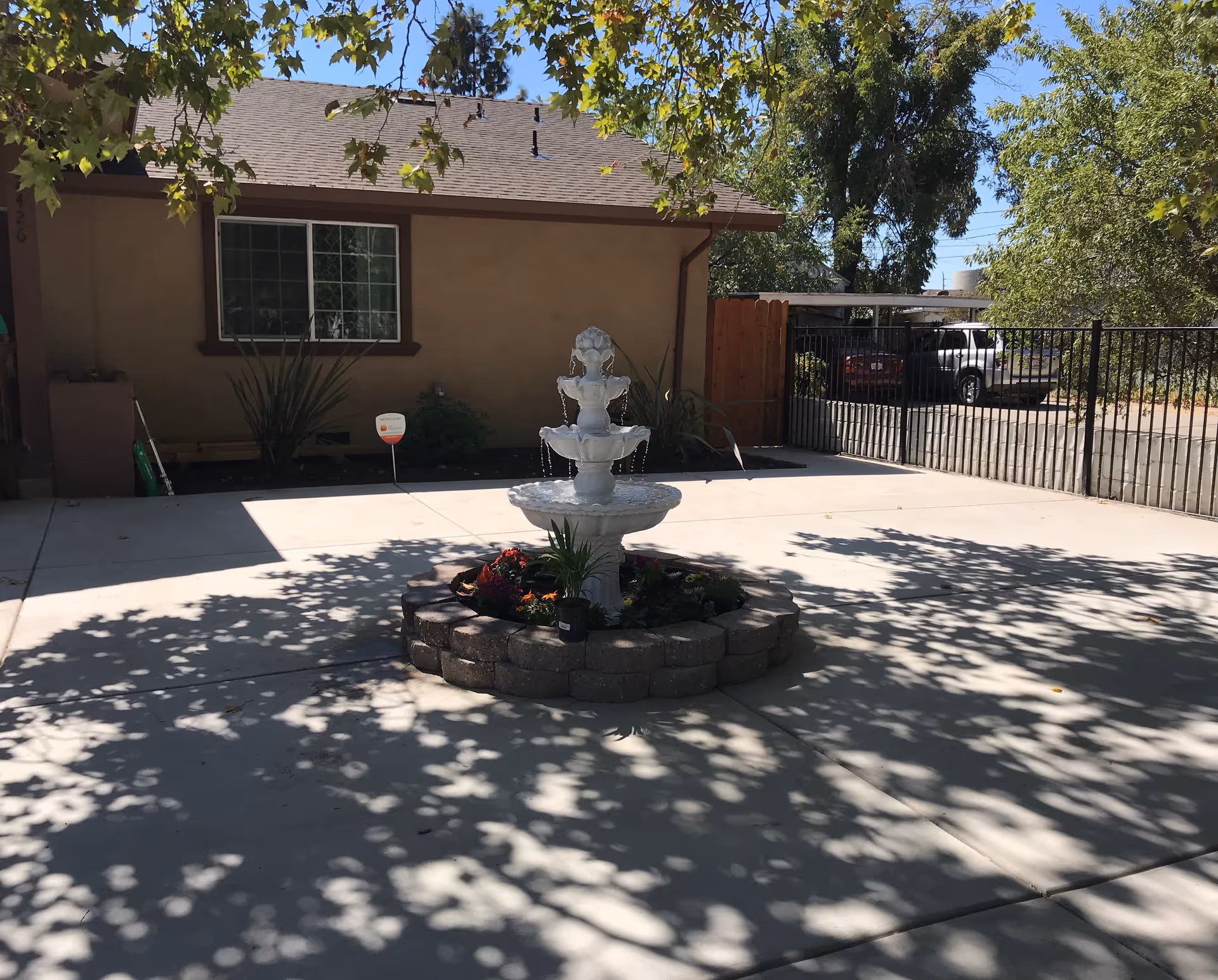 Outdoor courtyard area with a three-tier white water fountain surrounded by a circular stone planter with flowers. The courtyard is paved with concrete and shaded by tree branches. In the background, there is a beige building with a window and a black metal fence enclosing a parking area with cars.