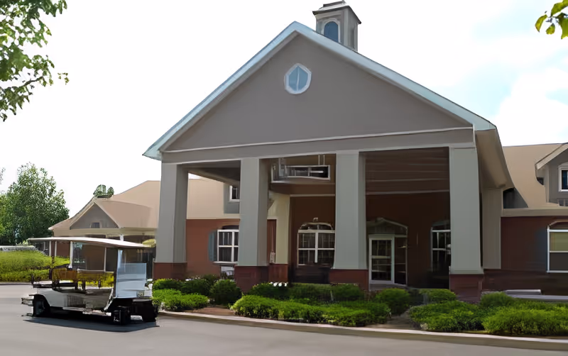 Main entrance of a senior living building with a large portico, landscaped shrubs, and a parked golf cart nearby.