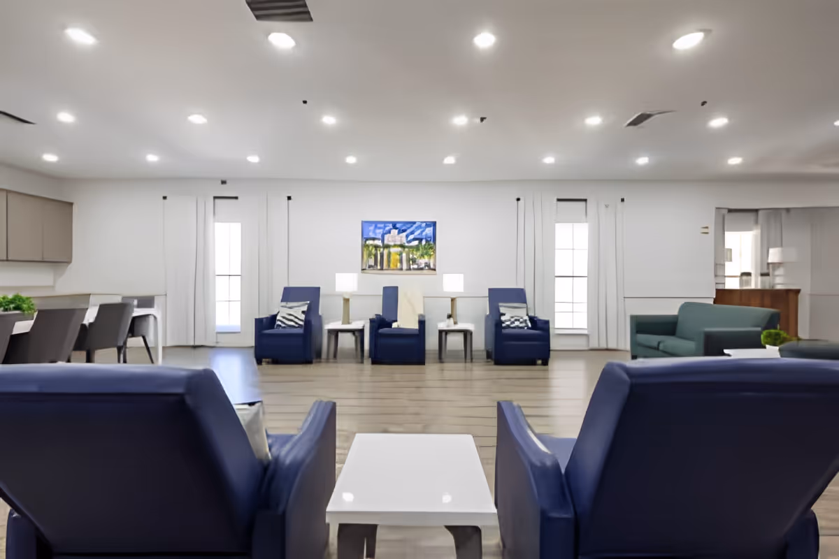 A spacious and well-lit common living area in a senior living facility with multiple blue armchairs and a green sofa arranged around white tables. The room has large windows with white curtains, wooden flooring, and a colorful painting on the wall. There are also lamps on side tables and a dining area with chairs visible on the left side.