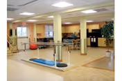 Interior view of a rehabilitation facility room with parallel bars for walking exercises, exercise equipment including a large red exercise ball, wooden flooring, and a kitchenette area in the background with cabinets and appliances.