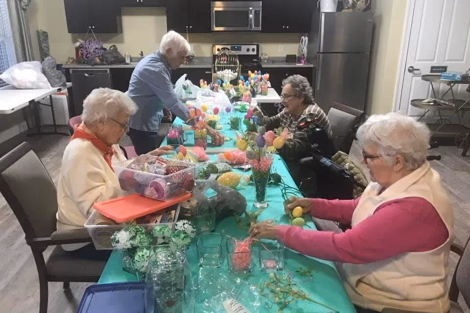 Four elderly women sitting around a long table in a kitchen area, engaging in a craft activity with colorful decorations and materials spread out on the table.