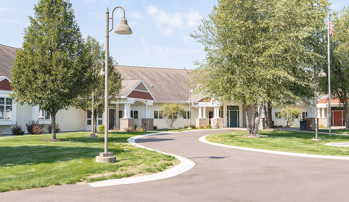 Exterior view of a senior living facility with a curved driveway, green lawns, several trees, and lamp posts. The building has a beige facade with white trim and a brown roof under a partly cloudy sky.