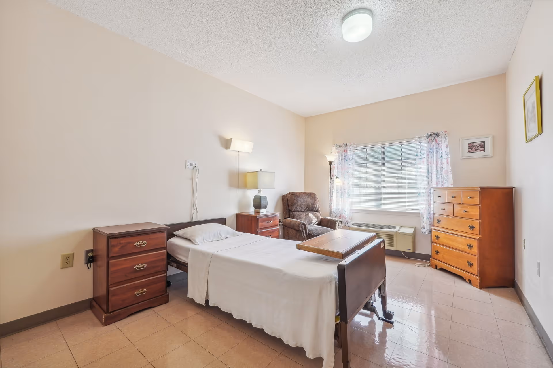 Furnished senior bedroom with a single bed, bedside tables, a reclining armchair and a dresser by a window.