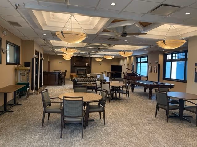 A spacious senior living common area with multiple round tables and chairs, a pool table, a grand piano, a fireplace, and large windows letting in natural light. The ceiling features recessed lighting and hanging light fixtures.