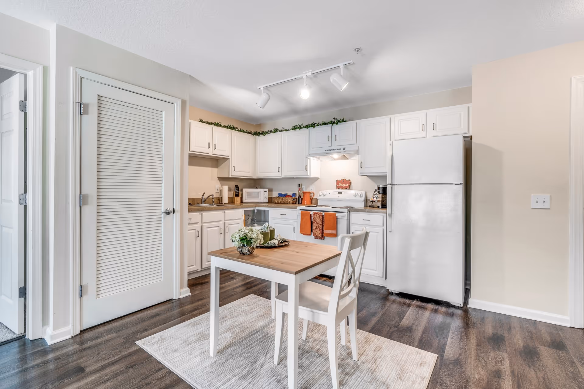 A bright kitchen area with white cabinets, a white refrigerator, a white stove with orange towels hanging on the handle, a microwave, and a small wooden table with a white chair on a light-colored rug. The kitchen has dark wood flooring and beige walls, with a door to the left and track lighting on the ceiling.