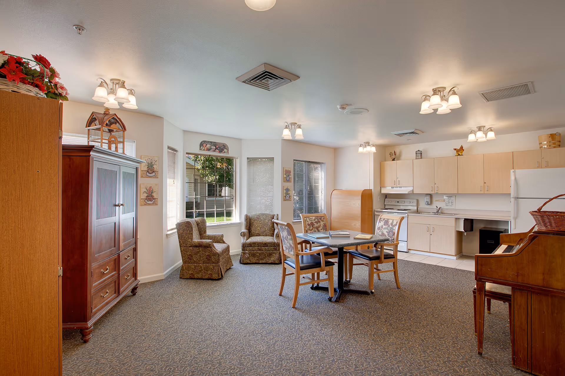 A bright and spacious common area in a senior living facility featuring a small kitchen with light wood cabinets, a white refrigerator, stove, and sink. In the foreground, there is a square table with four chairs around it, and two armchairs are placed near large windows that let in natural light. The room has carpeted flooring and multiple ceiling light fixtures. A wooden cabinet and a piano are also visible.