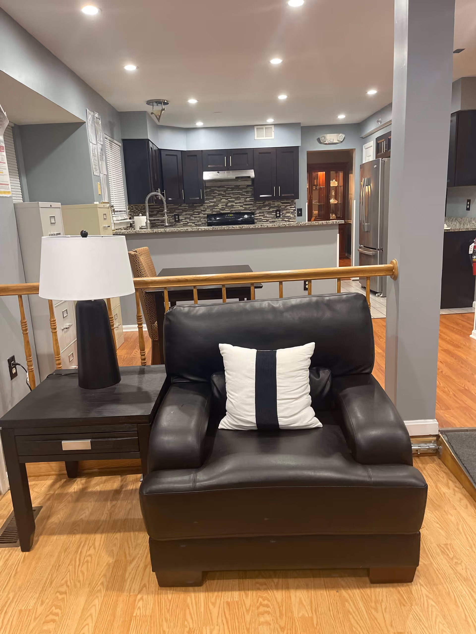 Black leather armchair with a white-striped pillow beside a side table and lamp in an open-plan living area facing a kitchen with dark cabinets and a breakfast bar.