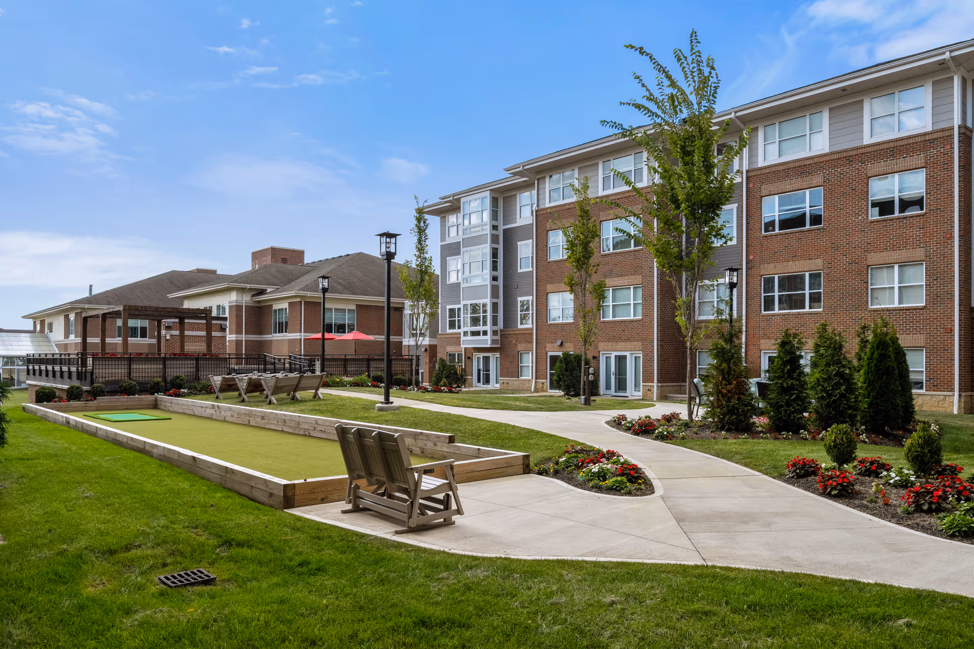 Outdoor view of Verena at Hilliard senior living facility showing a well-maintained lawn with a bocce ball court, wooden benches, paved walkways, flower beds, and a multi-story brick building under a clear blue sky.