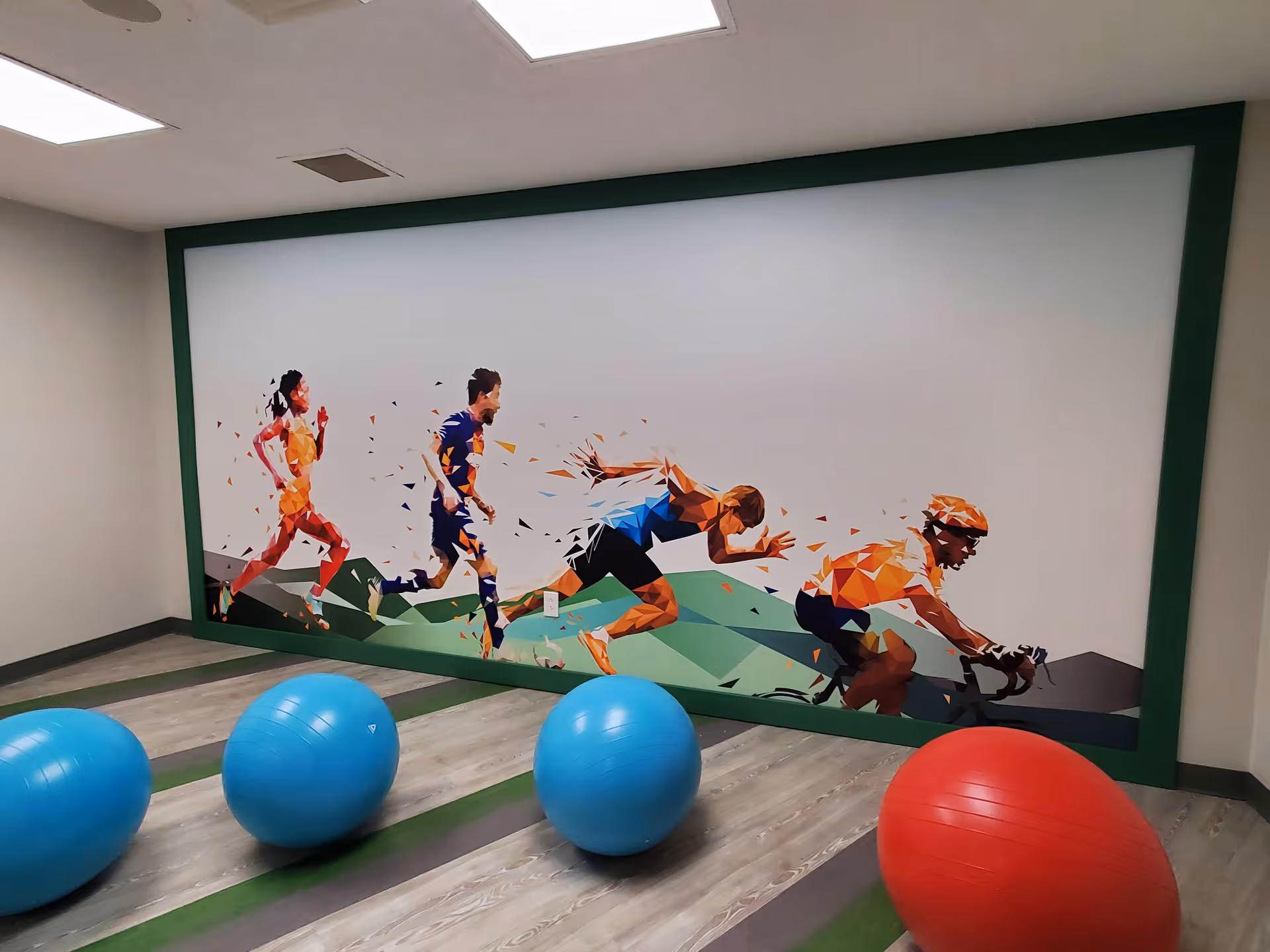 A fitness room with four exercise balls, three blue and one red, placed on a wood-patterned floor with green stripes. The back wall features a large colorful mural of four athletes in motion: a runner, a sprinter, a hurdler, and a cyclist, depicted in a geometric, fragmented art style.