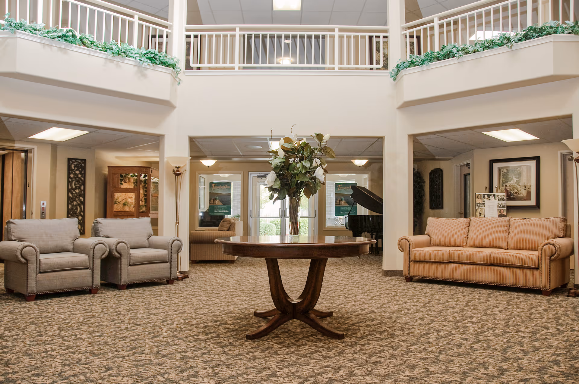 Bright senior living facility lobby with a round wooden table and floral centerpiece, sofas and armchairs beneath an upper balcony.