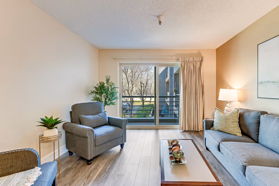 Cozy living room with a gray armchair and sofa, coffee table, lamp, plants, and a sliding glass door leading to a balcony.