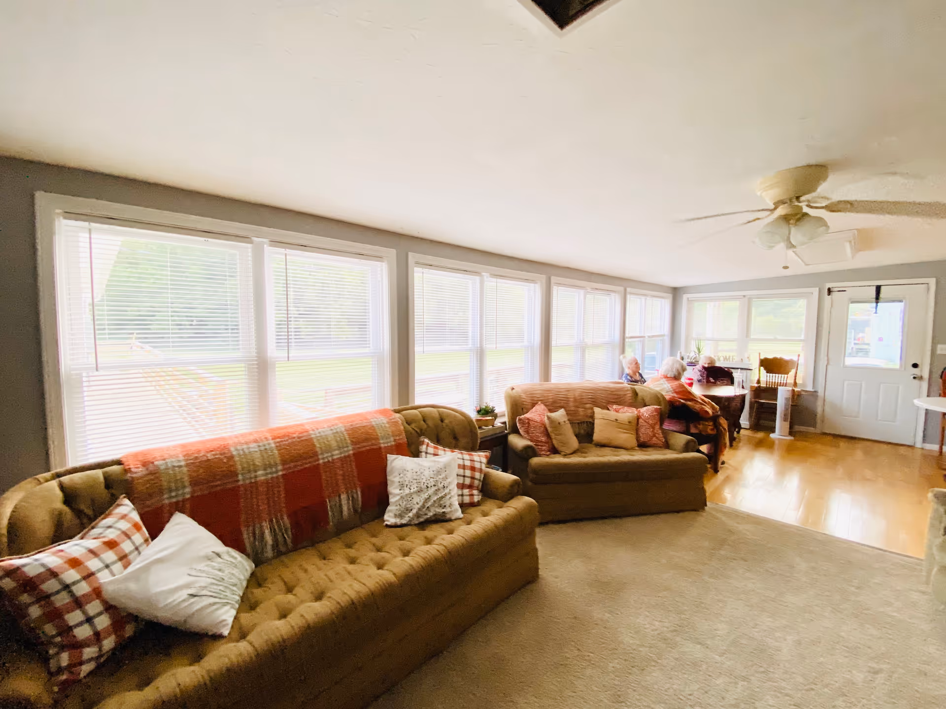 Sunlit living room with tufted sofas, throw pillows and blankets along a wall of large windows, with a small dining area and door at the far end.