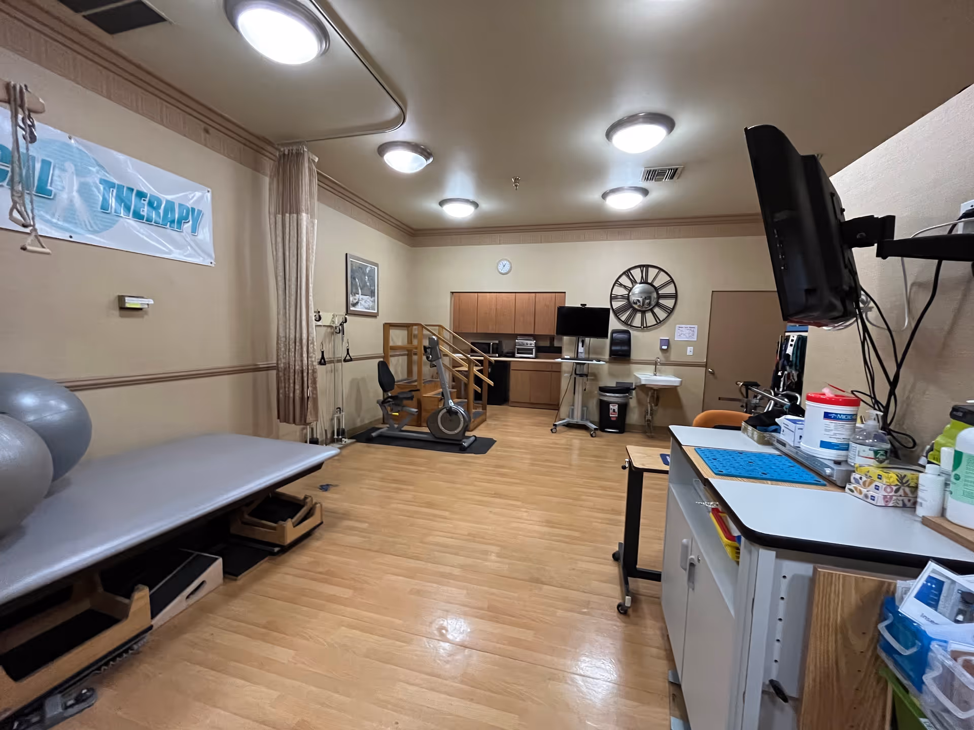 A physical therapy room with wooden flooring, exercise equipment including a stationary bike and therapy balls, a treatment table, a wall-mounted TV, cabinets, a sink, and a large decorative wall clock. The room is well-lit with ceiling lights and has a banner on the wall that reads 'THERAPY'.