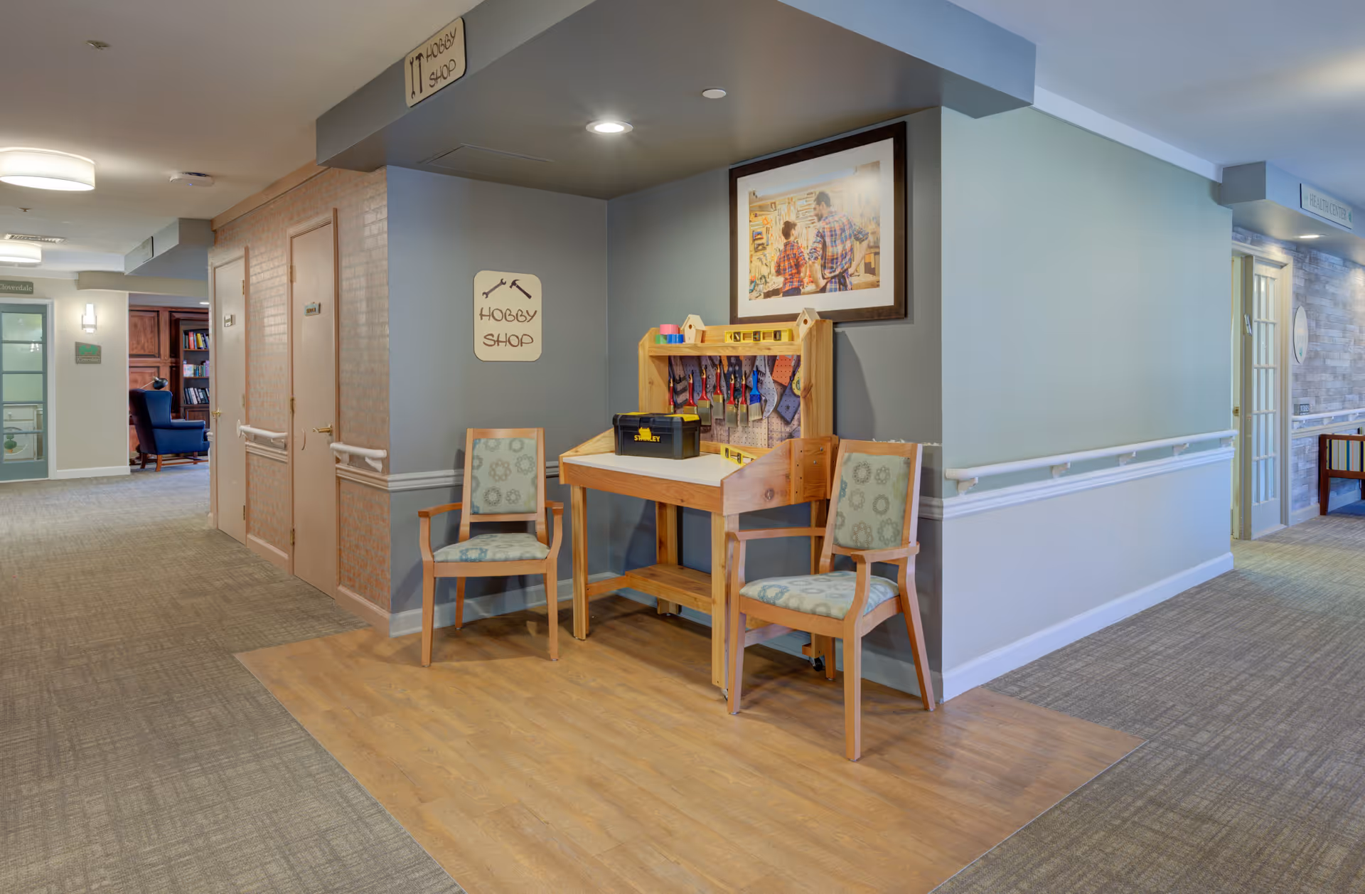Interior hallway of a memory care facility featuring a small hobby shop workstation with two chairs, tools, and framed artwork.