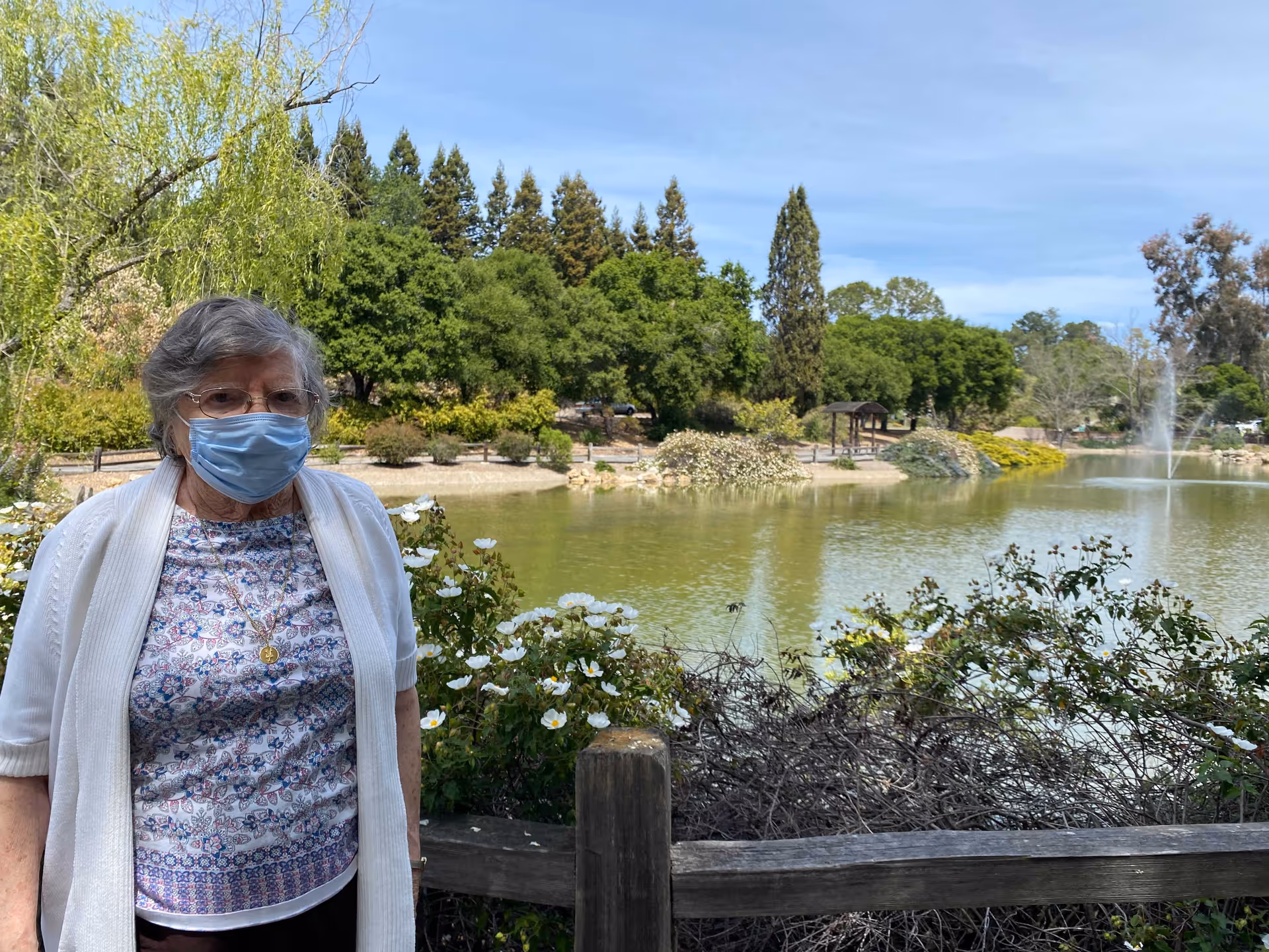 An elderly woman wearing a face mask, glasses, a patterned shirt, and a white cardigan stands near a wooden fence by a pond with white flowers and greenery. In the background, there are trees, bushes, and a water fountain in the pond under a clear blue sky.