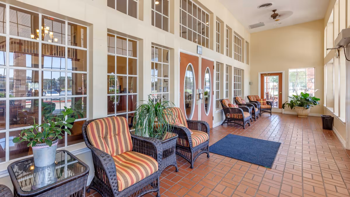 A bright indoor seating area with multiple wicker chairs featuring striped cushions arranged along a wall with large windows. There are potted plants on glass-top side tables and on the floor. The floor is tiled with reddish-brown tiles, and there are double doors with oval glass panels in the background.
