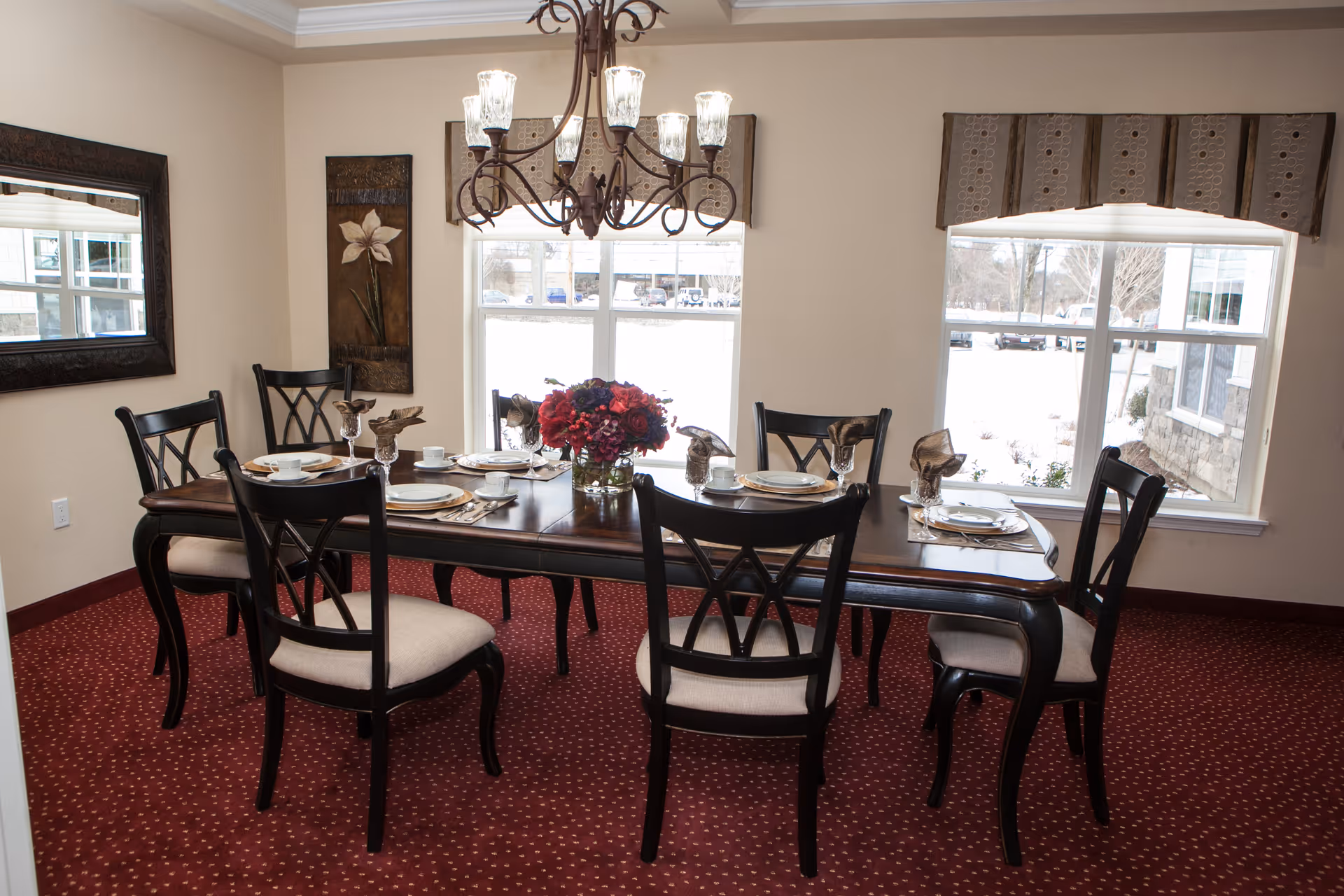 A dining room with a dark wooden table set for six people, featuring white plates, cups, and napkins folded in a decorative manner. The chairs have black frames with beige cushions. A chandelier with six lights hangs above the table. Two large windows with valances allow natural light to fill the room. The floor is covered with a red carpet with a small dotted pattern. A large mirror and a floral wall decoration are on the beige walls.