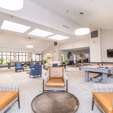 Spacious senior living facility common area with multiple seating arrangements including leather and fabric chairs around circular wooden tables. The room features large skylights, modern pendant lights, a pool table, and large windows allowing natural light. The decor is neutral with light-colored walls and carpeted floors.