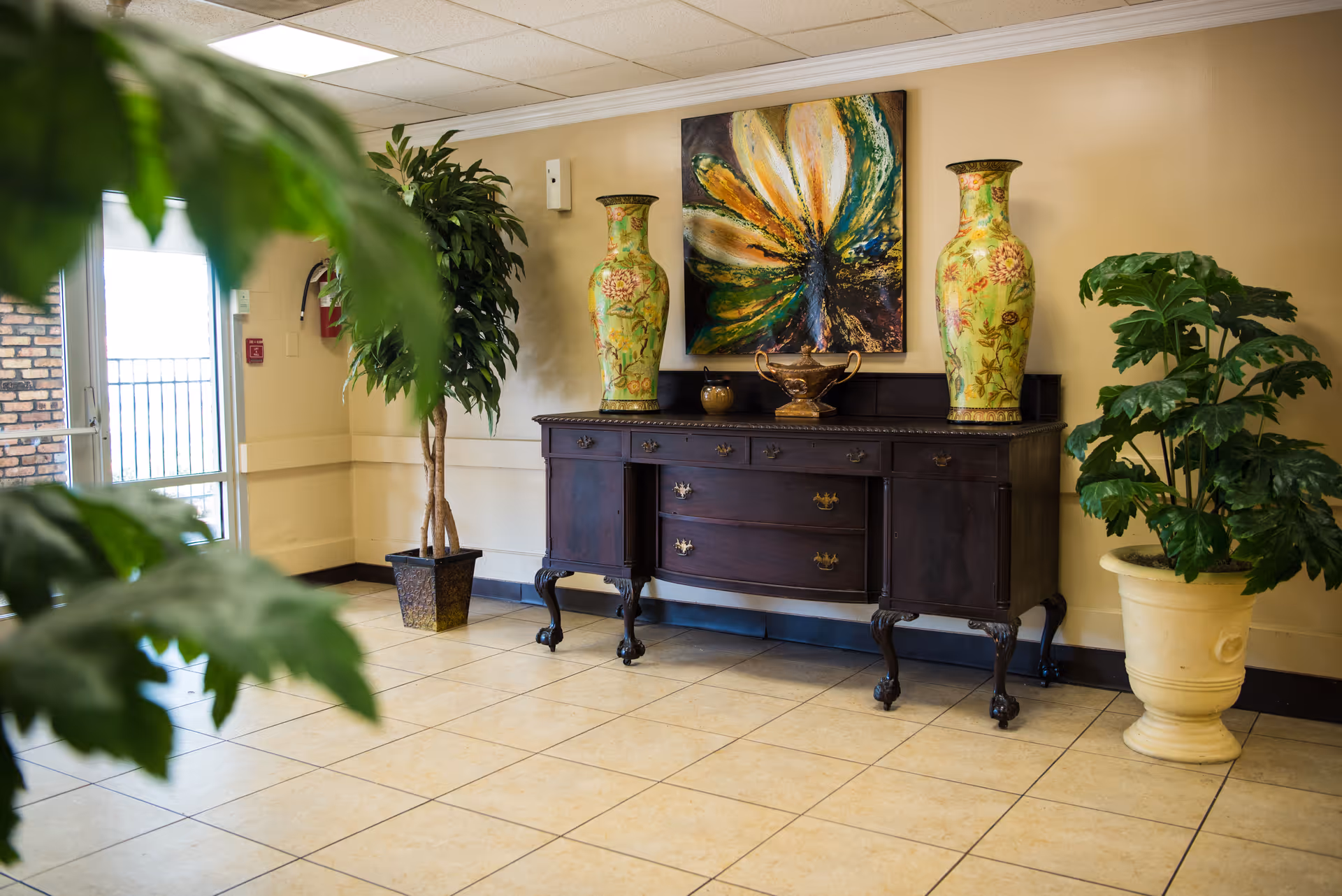 Lobby entryway with a dark wooden sideboard topped by two large decorative vases, a floral painting, and potted plants.