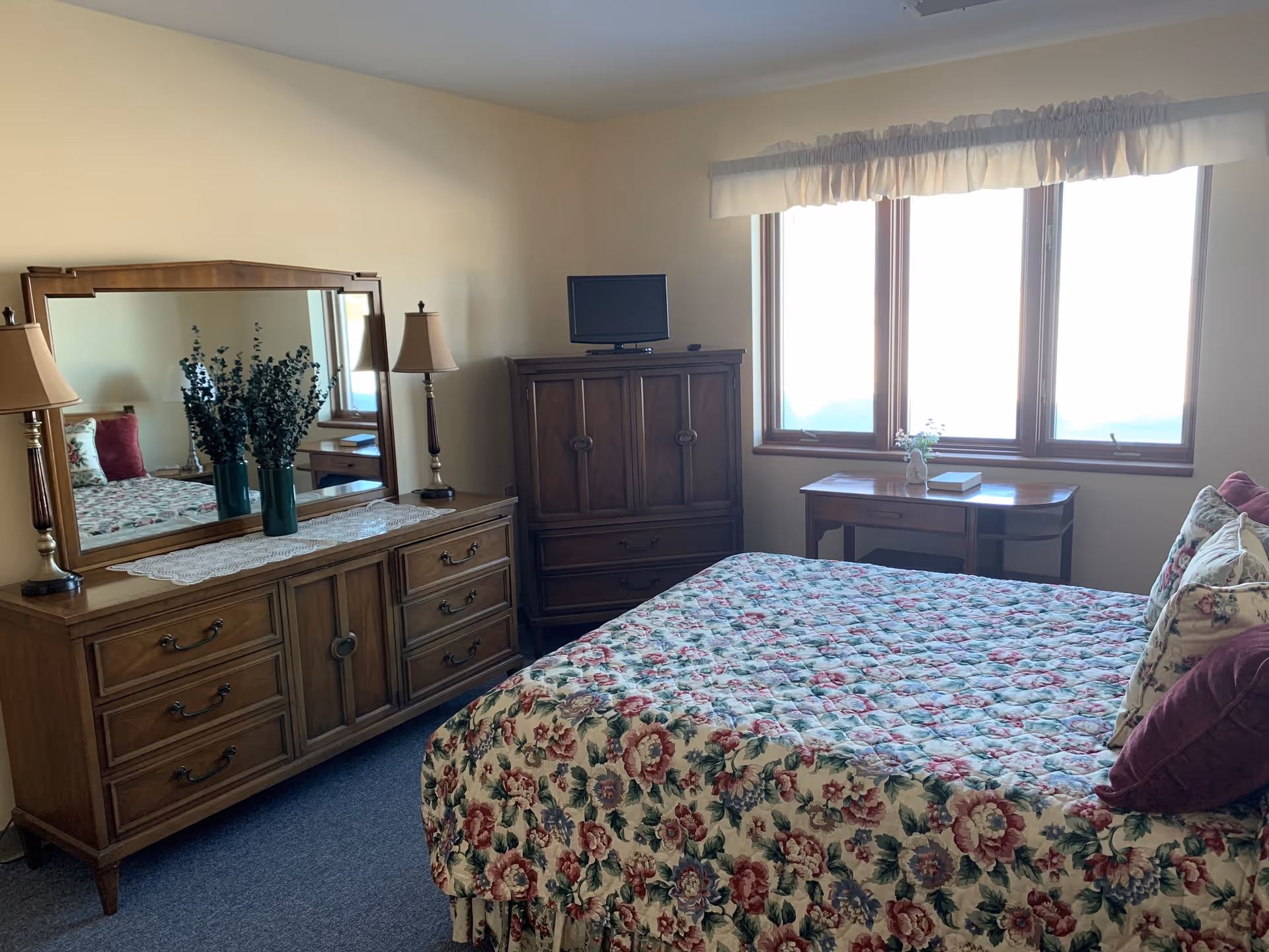 Bedroom with a floral quilted bed, wooden dresser with mirror and lamps, TV cabinet, and a windowed desk.