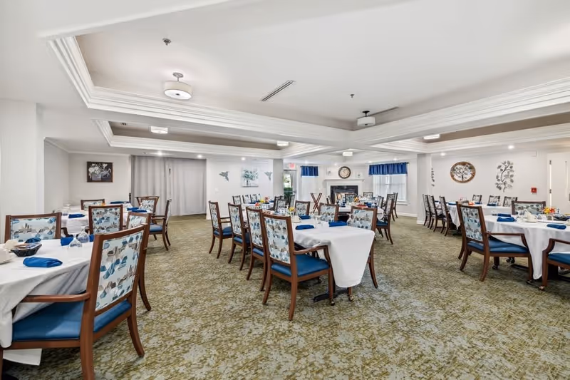 A spacious dining room with multiple tables covered in white tablecloths and set with blue napkins and glassware. The chairs have wooden frames with blue cushioned seats and floral-patterned backs. The room features a green patterned carpet, white walls, ceiling lights, and decorative wall art. There is a fireplace with a clock above it and windows with blue valances.