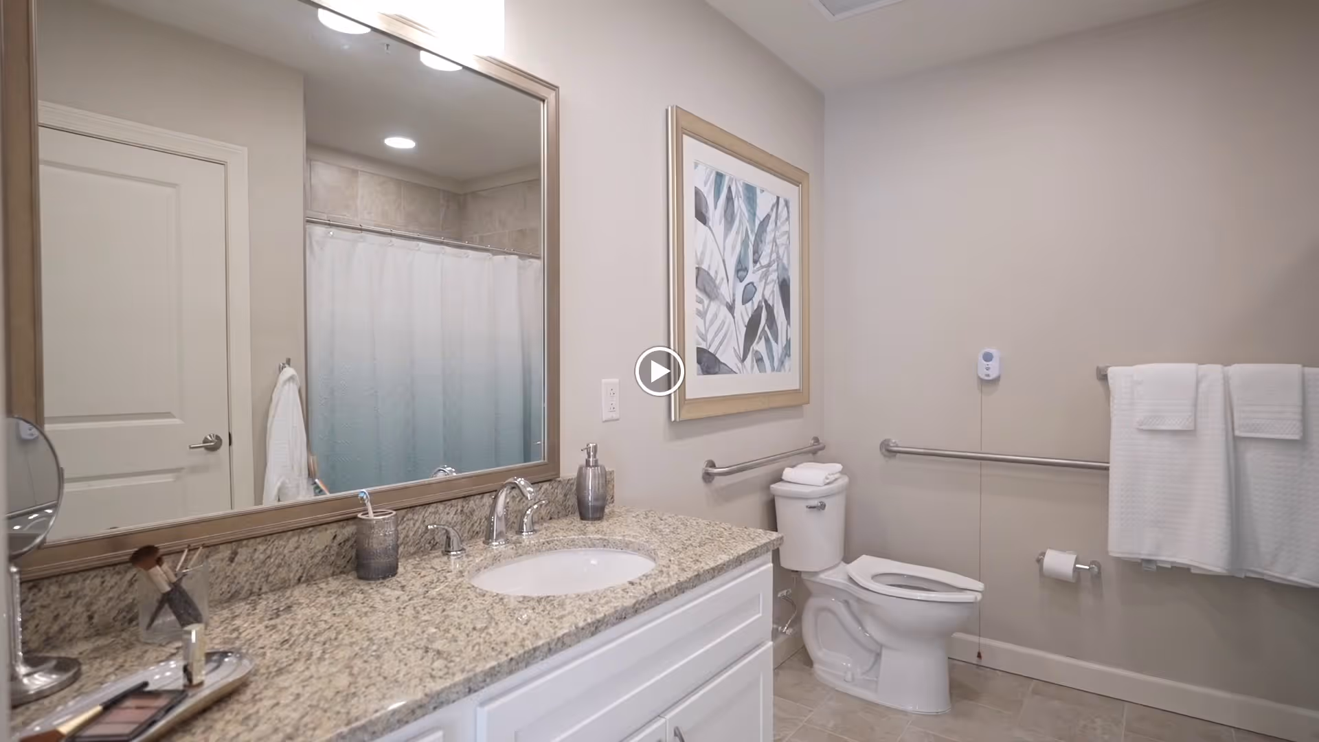 A clean and modern bathroom with a granite countertop, white sink, large mirror, toilet with grab bars, a towel rack with white towels, and a shower with a white curtain. There is a framed artwork on the wall above the toilet.